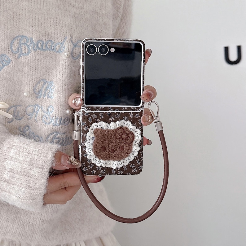 May include: A brown floral patterned phone case with a brown Hello Kitty appliqu&eacute; and a white lace trim. The case has a brown wrist strap and a silver-coloured clasp. The phone screen is black.