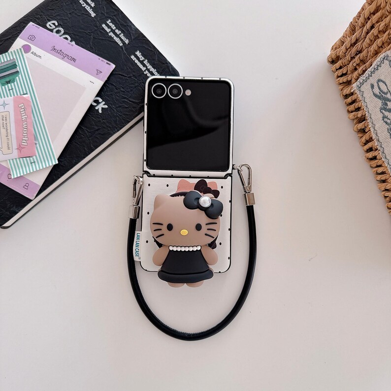 Op de afbeelding: Zwarte en witte smartphonehoes met een Hello Kitty-ontwerp. De hoes heeft een stippenpatroon en een zwarte band. De Hello Kitty-figuur draagt een zwarte jurk en een strik.