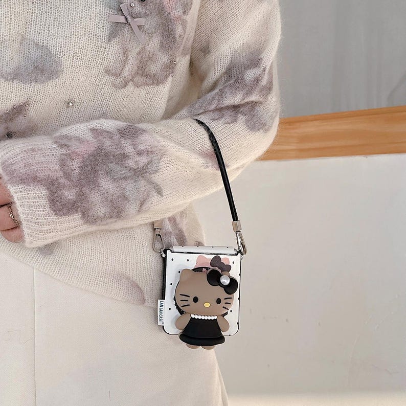 Op de afbeelding: Een kleine, vierkante crossbodytas met een witte achtergrond en zwarte stippen. Het heeft een 3D-cartoonfiguur met een zwarte strik en jurk. De tas heeft een zwarte band en zilverkleurige hardware.