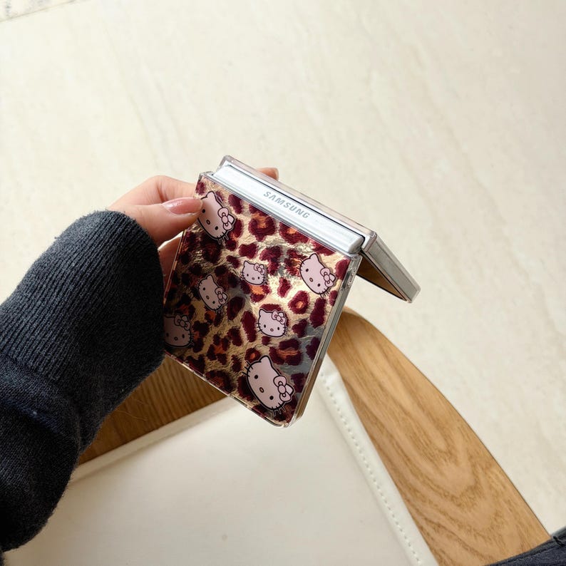 May include: A Samsung phone case with a leopard print pattern and Hello Kitty character illustrations. The case has a clear cover and a silver frame. The phone case is open, showing the phone's interior.