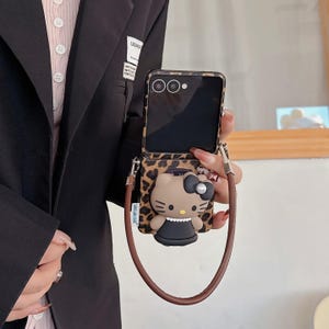 May include: A phone case with a leopard print design and a brown strap. The case features a 3D Hello Kitty character with a black bow and dress. The phone screen is visible.