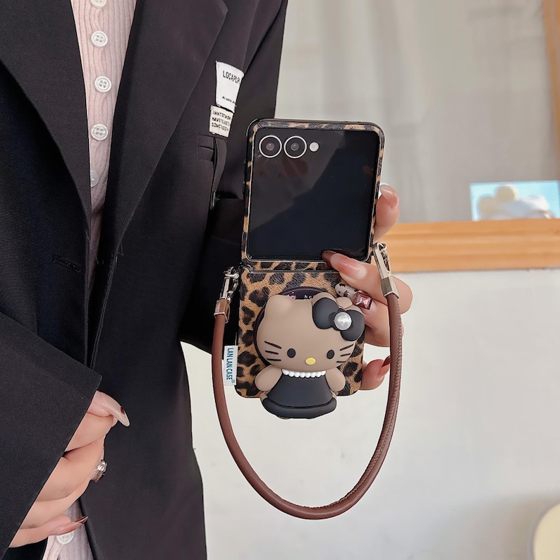 May include: A phone case with a leopard print design and a brown strap. The case features a 3D Hello Kitty character with a black bow and dress. The phone screen is visible.
