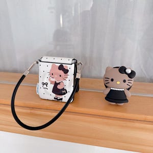 Op de afbeelding: Een witte AirPod-case met zwarte stippen, een zwarte band en een cartoonafbeelding van een kat met een zwarte jurk en een kopje. Er is ook een bijpassend kattenbeeldje in een zwarte jurk.