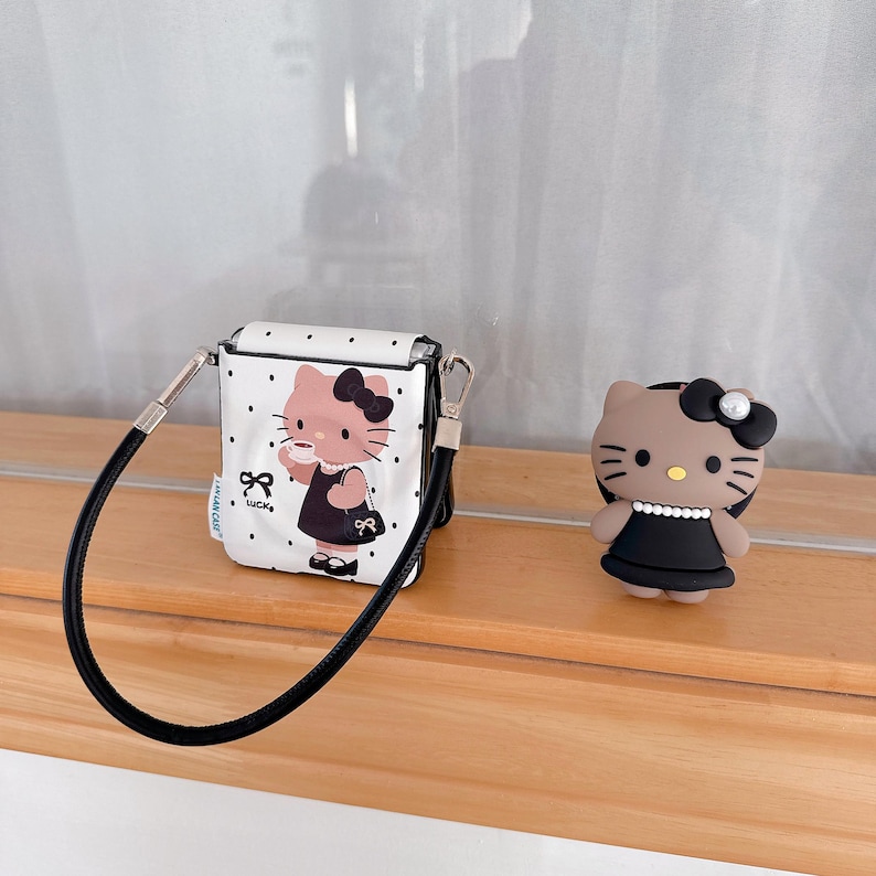 Op de afbeelding: Een witte AirPod-case met zwarte stippen, een zwarte band en een cartoonafbeelding van een kat met een zwarte jurk en een kopje. Er is ook een bijpassend kattenbeeldje in een zwarte jurk.