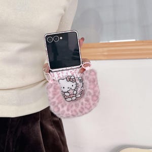 Op de afbeelding: Een roze en witte telefoonhoes met een Hello Kitty-ontwerp. De hoes heeft een fuzzy, luipaardprint zakje. De telefoon is zwart met een zilveren frame. De hoes wordt door een persoon vastgehouden.
