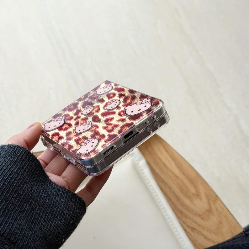May include: A clear phone case with a leopard print pattern in pink and brown. The case is decorated with pink Hello Kitty character designs. The case is held in a person's hand.