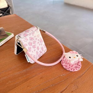 May include: A pink and white patterned phone case with a matching strap and a Hello Kitty charm. The case is open, revealing the phone's interior. The charm is a 3D Hello Kitty figure.