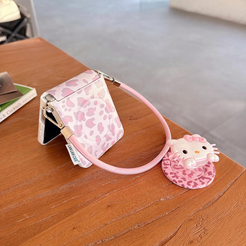 May include: A pink and white patterned phone case with a matching strap and a Hello Kitty charm. The case is open, revealing the phone's interior. The charm is a 3D Hello Kitty figure.