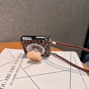 以下が含まれることがあります： ヒョウ柄のデザインの携帯電話ケース。小さな明るい色の漫画のキャラクターと茶色の革ストラップが付いています。ケースには「LAN LAN CASE」の文字が入っています。ケースはテキストのある白い紙の上にあります。