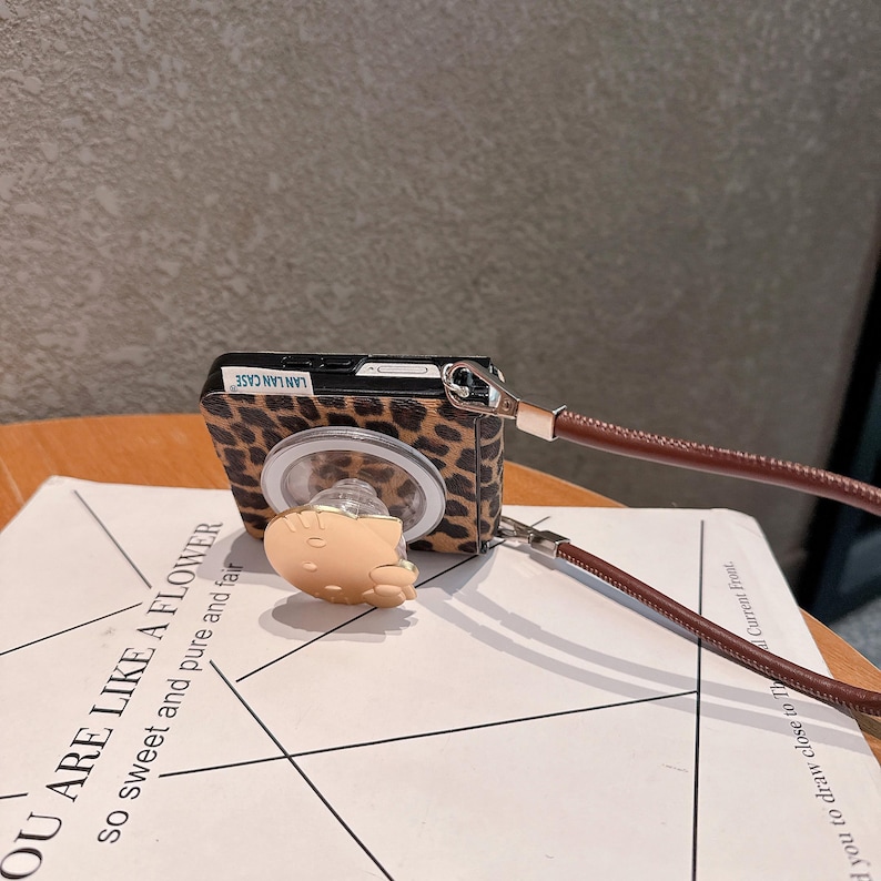 以下が含まれることがあります： ヒョウ柄のデザインの携帯電話ケース。小さな明るい色の漫画のキャラクターと茶色の革ストラップが付いています。ケースには「LAN LAN CASE」の文字が入っています。ケースはテキストのある白い紙の上にあります。