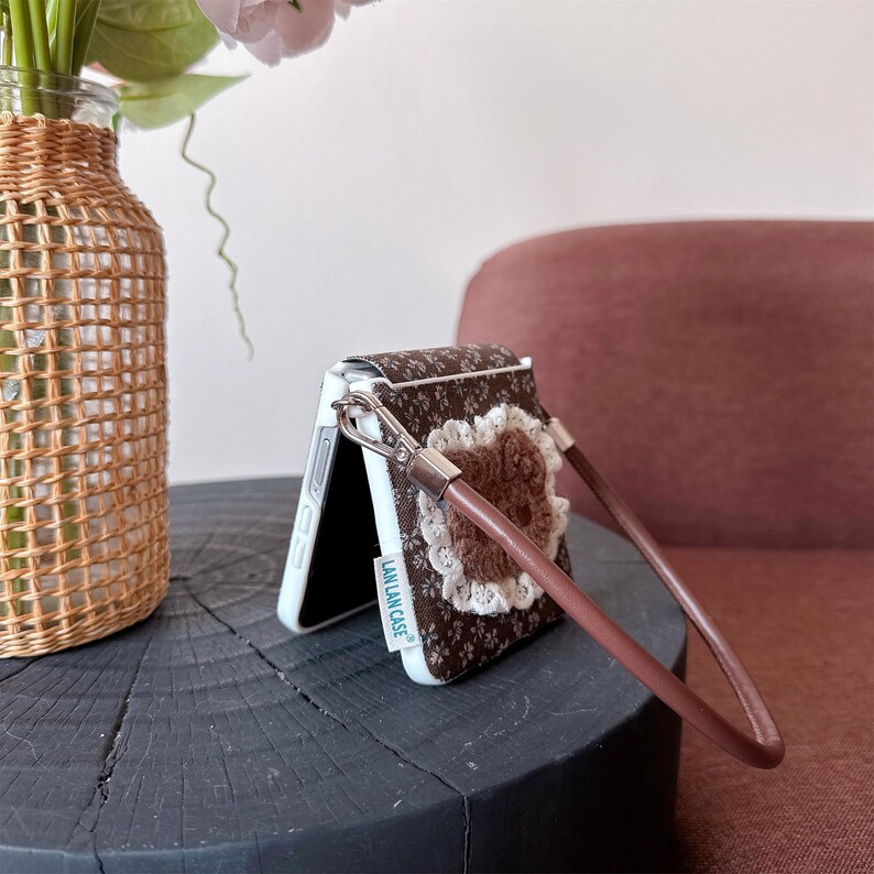 May include: A brown and white floral patterned phone case with a brown leather wrist strap. The case features a crocheted flower and white lace trim. The phone case is open, revealing a white interior. The text "LAN LAN CASE" is visible.