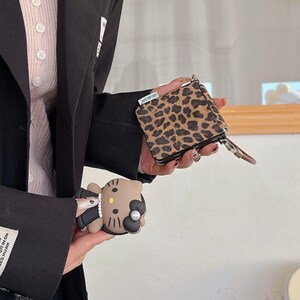 May include: A small, leopard print wallet with a zipper and a brown leather pull tab. A brown and black Hello Kitty phone case is also visible. The items are held against a neutral background.