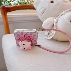 May include: A pink and white phone case with a camera design. The case has a pink leopard print pattern and a 3D Hello Kitty figure. It includes a pink strap and the text "LAN LAN CASE".