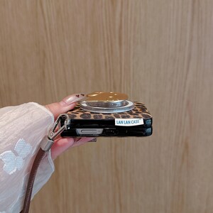 以下が含まれることがあります： ヒョウ柄の携帯電話ケースとゴールドカラーのポップアップグリップ。ケースには茶色のリストストラップと「LAN LAN CASE」の文字が入っています。
