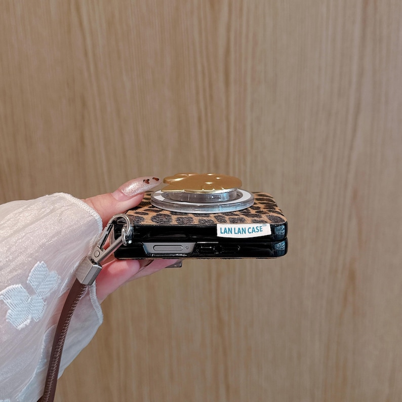以下が含まれることがあります： ヒョウ柄の携帯電話ケースとゴールドカラーのポップアップグリップ。ケースには茶色のリストストラップと「LAN LAN CASE」の文字が入っています。