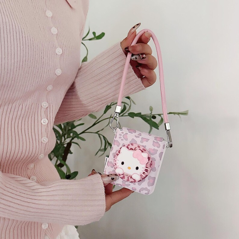 May include: A pink and white square crossbody bag featuring a Hello Kitty design. The bag has a pink strap and a leopard print pattern. The Hello Kitty design is in the center of the bag.