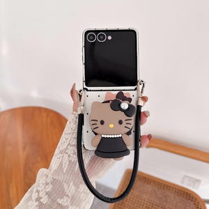 Op de afbeelding: Een telefoonhoesje met een cartoon kattenontwerp. De hoes is wit met zwarte stippen en een zwarte band. De kat heeft een zwarte strik, een parel en een zwarte jurk. De telefoon is zwart.