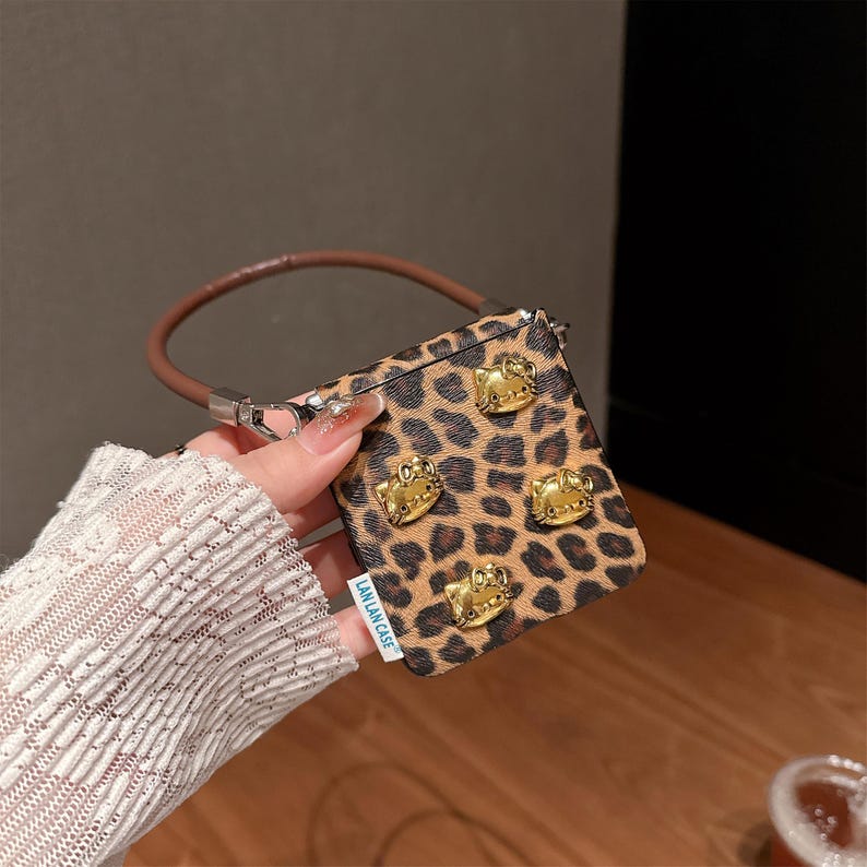 May include: A small, square handbag with a brown and black leopard print pattern. The bag features four gold-colored cat-shaped embellishments and a brown leather handle. The text "LAN LAN CASE" is visible on the side.