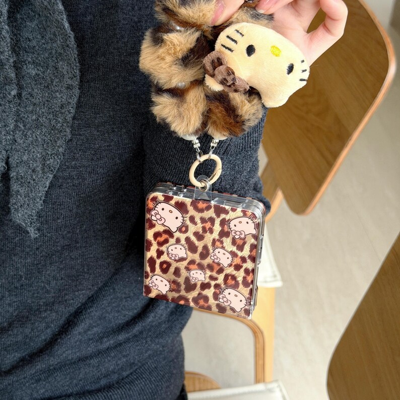 May include: A clear, square phone case featuring a leopard print pattern and Hello Kitty motifs. It's connected to a keychain with a fluffy leopard print charm and a small Hello Kitty plush toy.