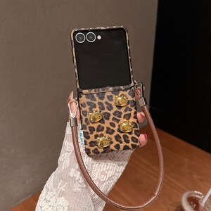 May include: A leopard print phone case with gold-colored Hello Kitty embellishments. The case has a brown leather strap and a black phone screen. The case is designed to hold a phone.