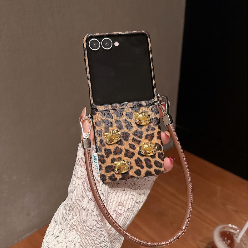 May include: A leopard print phone case with gold-colored Hello Kitty embellishments. The case has a brown leather strap and a black phone screen. The case is designed to hold a phone.