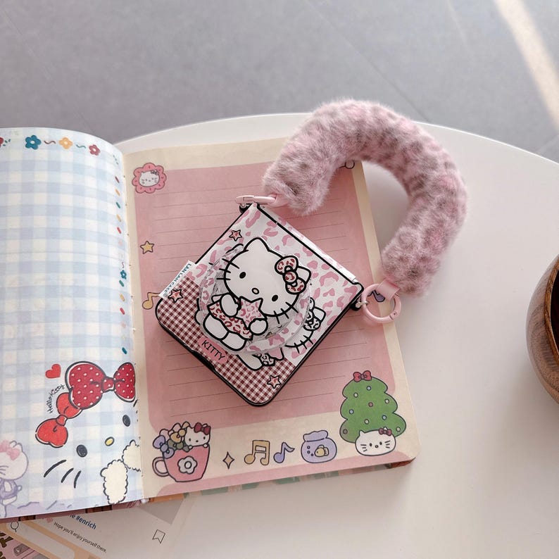 Op de afbeelding: Een roze en witte Hello Kitty-hoes met een donzige, lichtroze polsband. De hoes heeft een cartoonafbeelding van Hello Kitty en staat op een roze notitieboekpagina met andere Hello Kitty-illustraties.