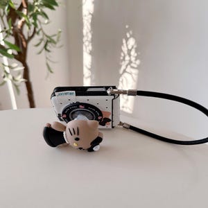 Op de afbeelding: Een wit en zwart telefoonhoesje met een camera-ontwerp en een zwarte band. De hoes heeft een stripfiguur en de tekst "LAN LAN CASE". Een bijpassende stripfiguur telefoonhouder is bevestigd.