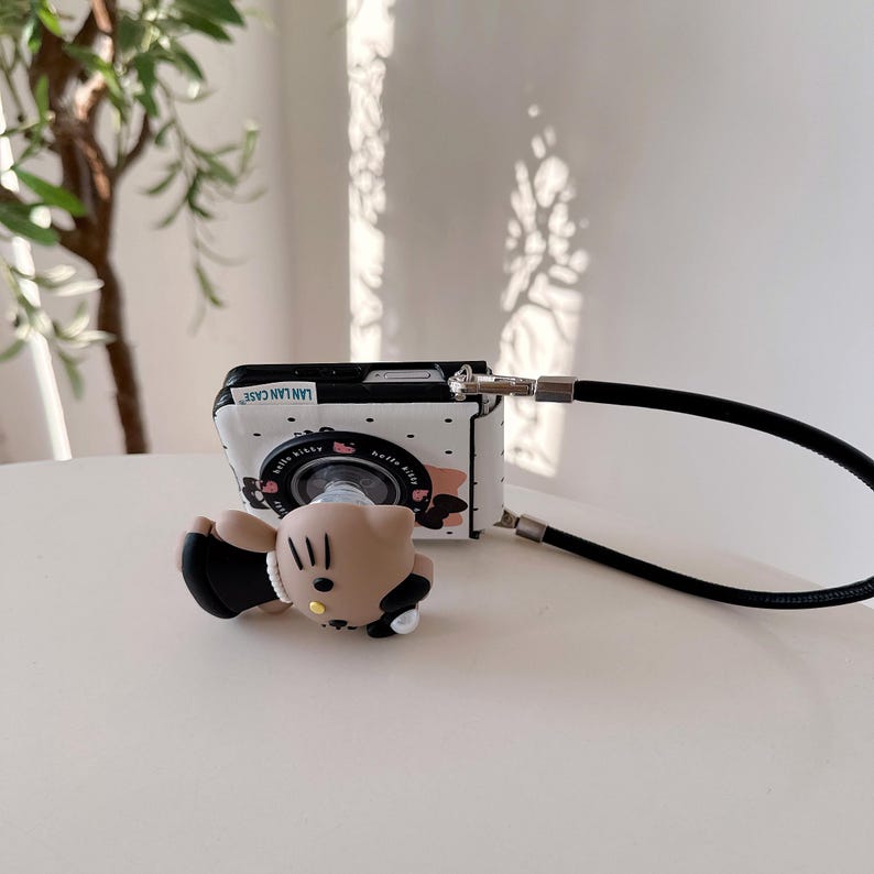 Op de afbeelding: Een wit en zwart telefoonhoesje met een camera-ontwerp en een zwarte band. De hoes heeft een stripfiguur en de tekst "LAN LAN CASE". Een bijpassende stripfiguur telefoonhouder is bevestigd.