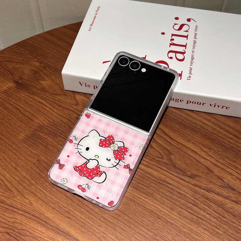 May include: A clear phone case featuring a pink and white checkered pattern with a Hello Kitty graphic. The phone case is open, revealing a black screen. The design includes cherries and bows.