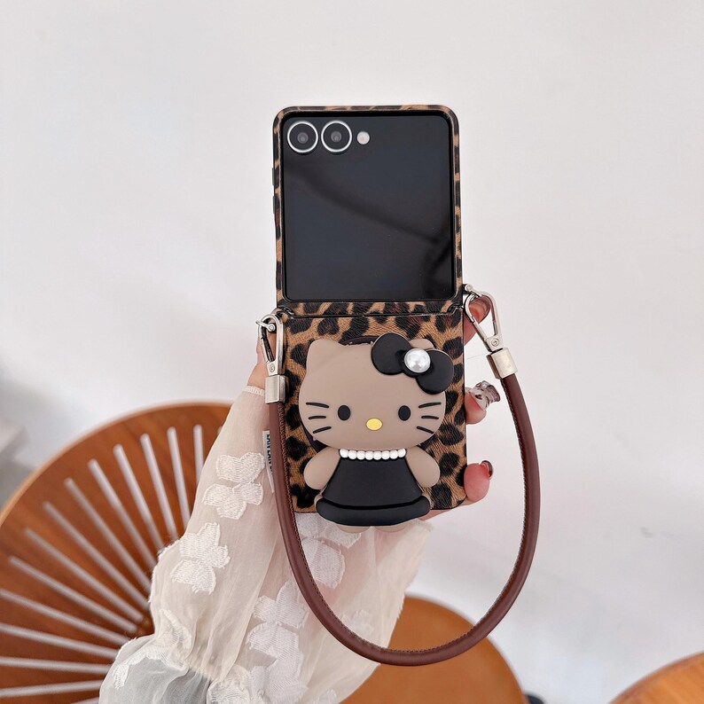 May include: A phone case featuring a leopard print design and a 3D cartoon character. The case has a brown strap and a black phone screen. The cartoon character has a black bow and a black dress.