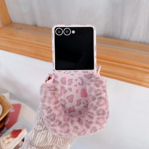 Op de afbeelding: Een roze en grijze telefoonhoes met luipaardprint en een pluizige, imitatiebonten band. De telefoonhoes is open en onthult een zwart scherm en cameralenzen. Ontworpen voor een opvouwbare telefoon.