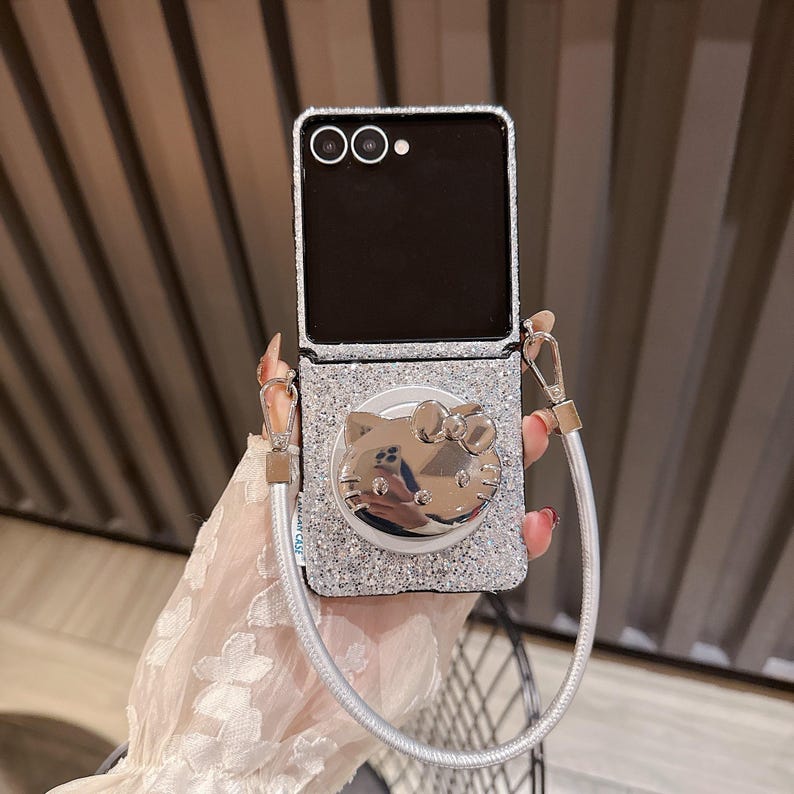 May include: Silver glitter phone case with a silver Hello Kitty design and a matching silver strap. The phone case is open, revealing a black screen. The case has a silver glitter finish and a silver pop-up grip.