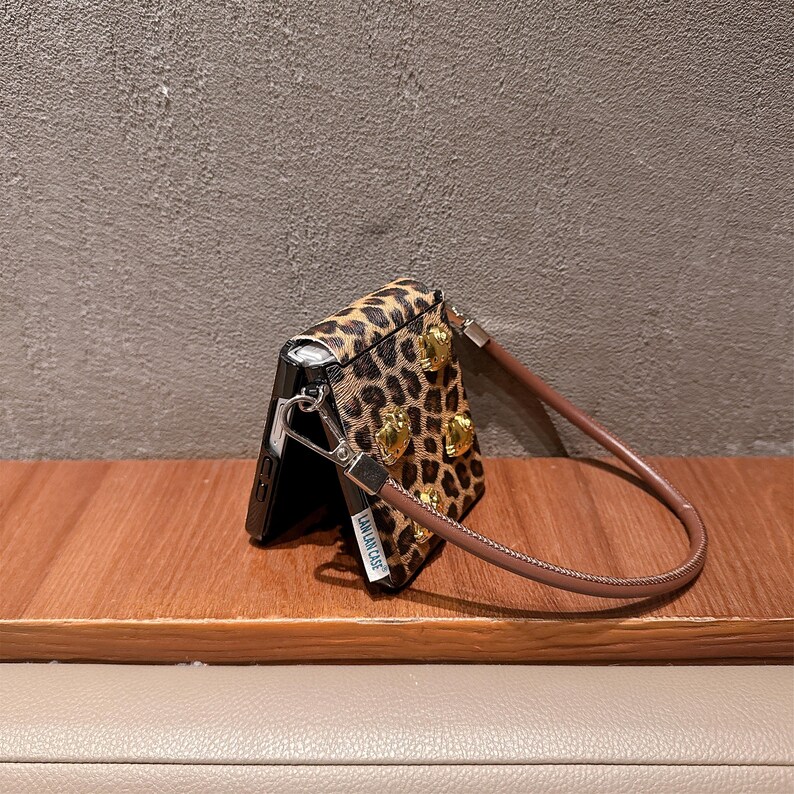 May include: A leopard print phone case with gold-colored heart-shaped embellishments and a brown wrist strap. The case is open, revealing the phone's screen. The text "LALA LAN CASE" is visible on the side of the case.