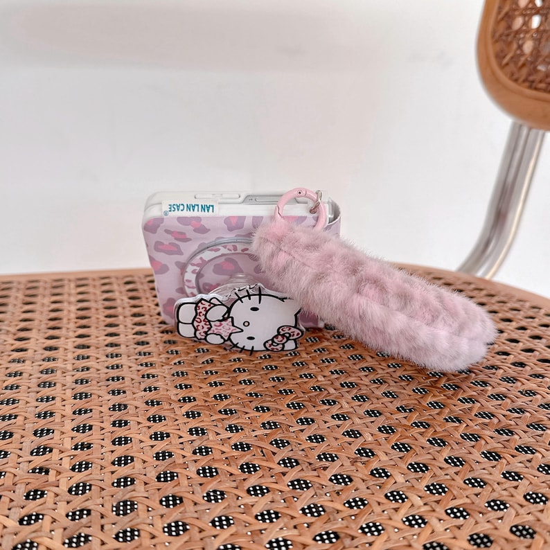 Op de afbeelding: Een verzameling roze accessoires. Inbegrepen zijn een doorzichtige telefoonhoes met een roze luipaardprint, een roze fluffy sleutelhanger en een Hello Kitty-sticker. De telefoonhoes heeft de tekst "LAN LAN CASE".