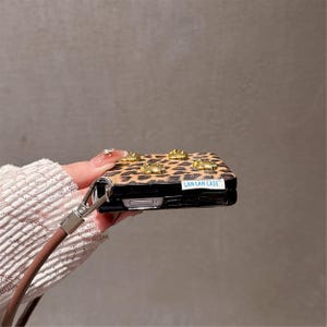 May include: A leopard print phone case with gold-colored embellishments. The case has a brown strap and the text "LAN LAN CASE".