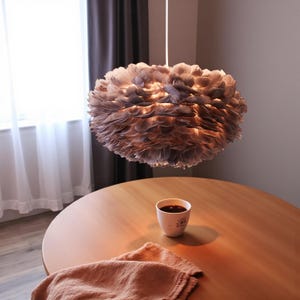 May include: A large, round pendant light made of layered, light-coloured feathers hangs from the ceiling. The light is on, illuminating the feathers. A wooden table with a cup of coffee and a cloth is in the foreground.