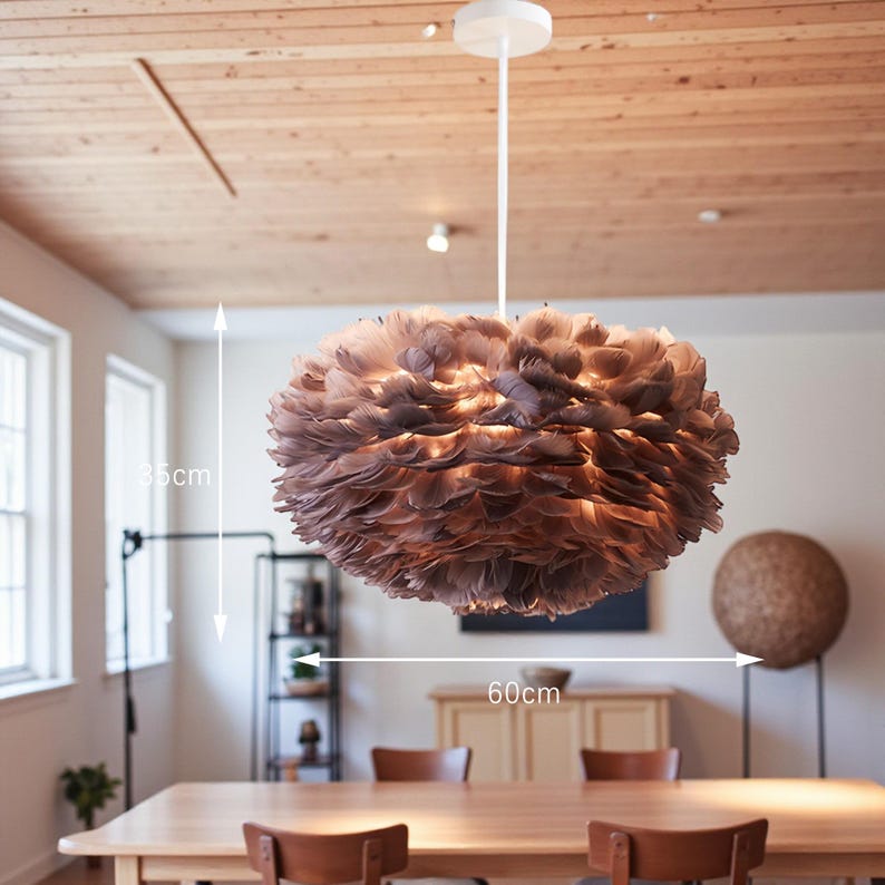 May include: A pendant light with a round, layered shade made of brown feathers. The shade is 60cm wide and 35cm high. The fixture hangs from a white ceiling mount and cord. The light is suspended above a wooden dining table.