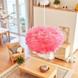 May include: A pink feather pendant light, 60 cm in diameter and 35 cm in height, suspended from the ceiling. The light fitting is round and fluffy, with a white ceiling mount and stem. The light is in a living room setting.