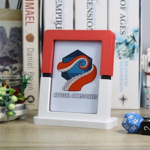 May include: A white and red tabletop display stand with a card featuring a blue and orange octopus design and the text "ABYSSAL ACCESSORIES." A blue and black die are in front of the stand.