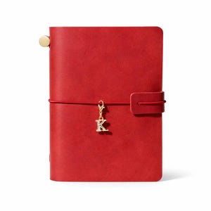 May include: A red leather journal with a gold-colored charm in the shape of the letter "K". The journal has a matching red elastic closure and a gold-colored button on the spine. The journal is a rectangular shape.