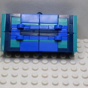 Puede incluir: Un vehículo de juguete de construcción rectangular hecho de ladrillos de plástico entrelazados en tonos azul oscuro, azul medio y verde azulado. La parte inferior presenta cuatro ruedas pequeñas azules unidas al chasis central, con un diseño plano y simétrico.