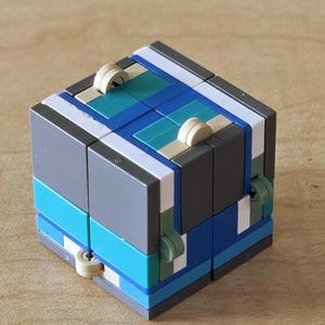 May include: A multi-coloured cube puzzle featuring shades of blue, grey, white, and teal. The cube has a complex, layered design with small, circular knobs. The puzzle is made of plastic and rests on a wooden surface. The cube measures approximately 5 cm x 5 cm.
