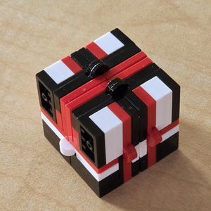 May include: A geometric puzzle cube in a bold colour scheme. The cube has alternating black, white, and red sections, with small black circular accents. The design is symmetrical, creating a visually engaging and complex structure. The cube is a brain teaser.