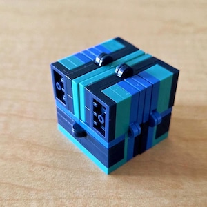 Puede incluir: Un rompecabezas de cubo multicolor con bloques azules, turquesas y negros. El cubo está ensamblado con piezas entrelazadas y tiene un diseño complejo. El cubo está sobre una superficie marrón claro.