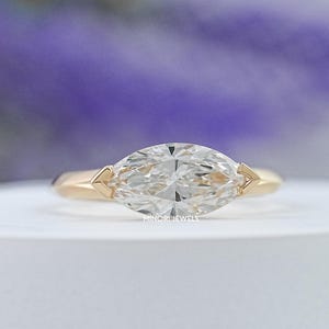 May include: A gold engagement ring with a marquise-cut diamond. The ring features a slender band and delicate side accents. The diamond is set horizontally, creating a unique and elegant design. The ring is displayed on a white surface with a blurred purple background.