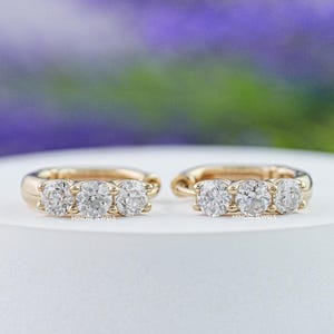 Puede incluir: Dos pendientes de aro dorados, cada uno con tres piedras preciosas redondas y transparentes. Los pendientes se exhiben sobre una superficie blanca, con un fondo borroso de color púrpura y verde.