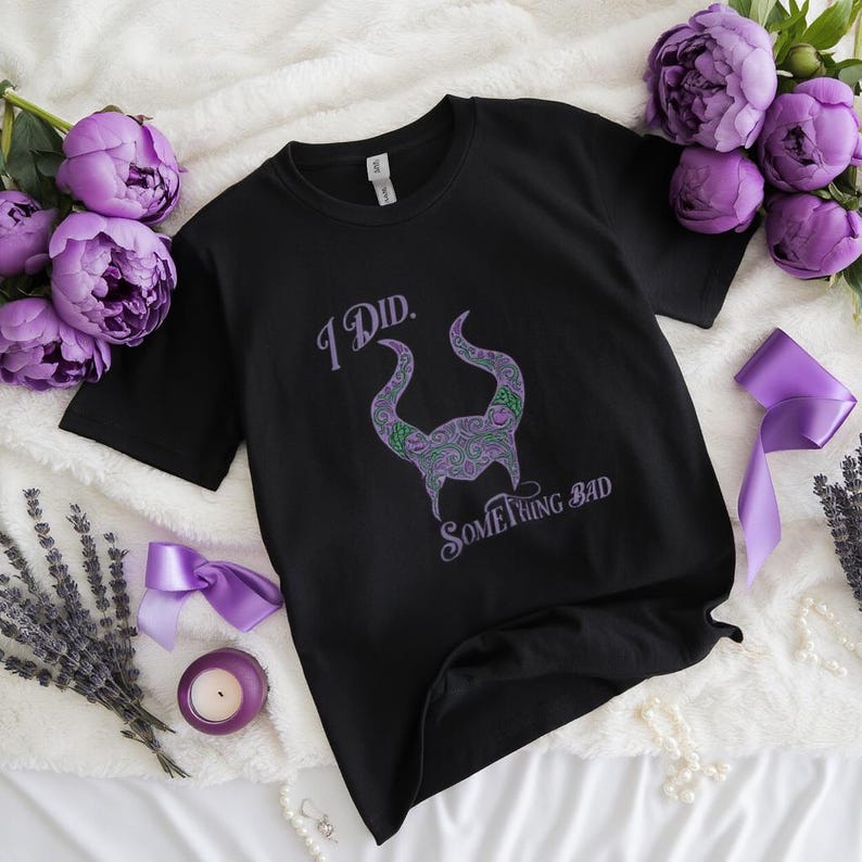 May include: Black t-shirt with the text "I DID. SOMETHING BAD" in purple script. The shirt features a purple and green graphic of a horned silhouette. The shirt is displayed on a white, textured surface with purple flowers and ribbons.