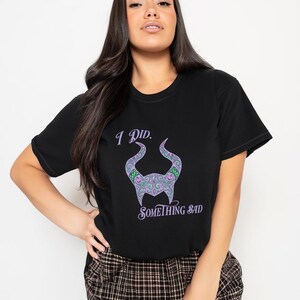May include: Black t-shirt with the text "I Did. Something Bad" and a purple and green horned design. The shirt is a crew neck and has short sleeves. The model is wearing a plaid skirt.