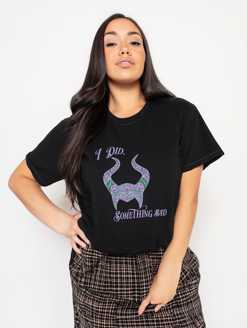May include: Black t-shirt with the text "I Did. Something Bad" and a purple and green horned design. The shirt is a crew neck and has short sleeves. The model is wearing a plaid skirt.