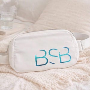 Puede incluir: Riñonera blanca con cremallera plateada y correa ajustable a juego. La bolsa presenta las iniciales "BSB" en azul turquesa. La bolsa está sobre una manta blanca.
